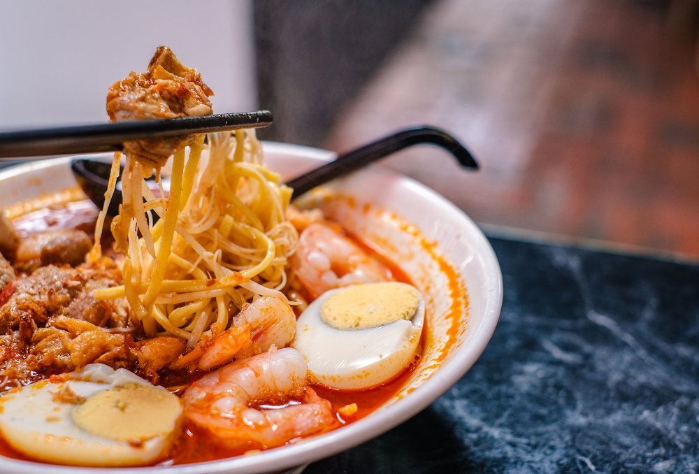 Chopsticks lifting noodles from a soup with eggs and prawns.