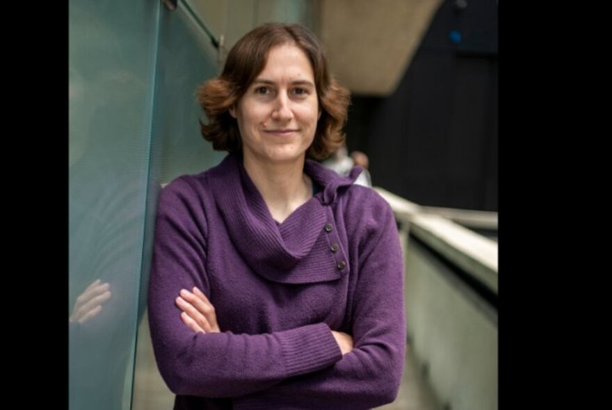 Katie Mack, a woman with shoulder length brown hair, her arms crossed in front of her chest, leaning against a wall.