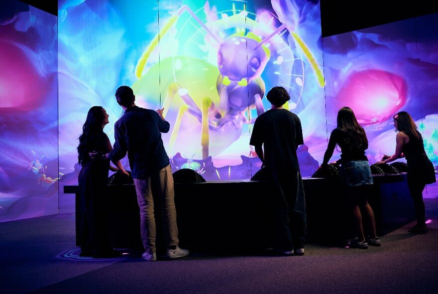 Silhouettes of people looking at giant projections of ants in a room with screens, the images are swirly and blue and purple tones. 