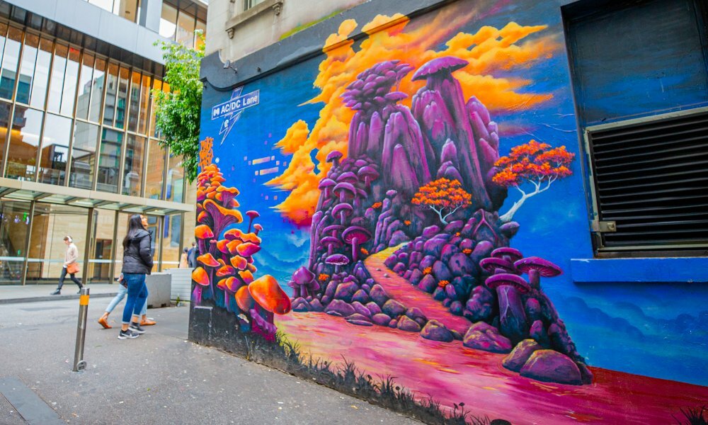 People walking past a mural of a purple mountain with orange mushrooms and clouds.