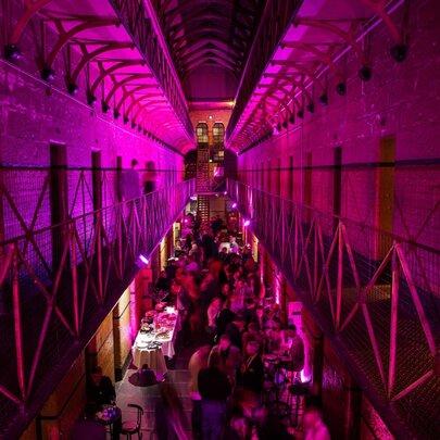 An event setup inside the historic cell block of the Old Melbourne Gaol showing a view of the ground floor with people socialising, as seen from the balcony level of the cell block.