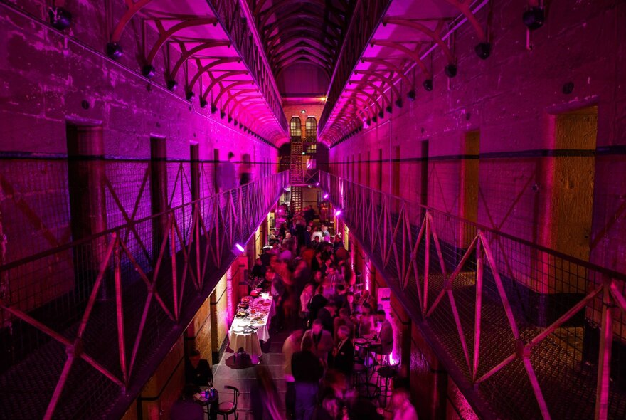 An event setup inside the historic cell block of the Old Melbourne Gaol showing a view of the ground floor with people socialising, as seen from the balcony level of the cell block.