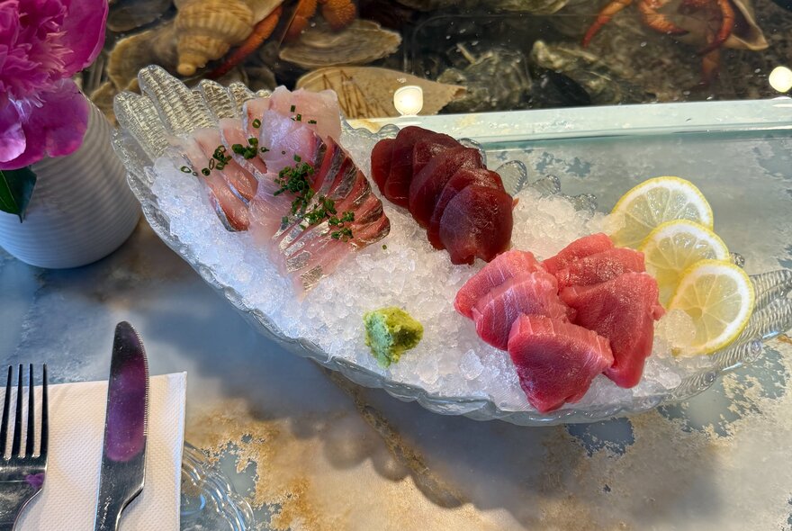 A selection of sashimi resting on a bed of ice in a glass serving dish on a marble table.