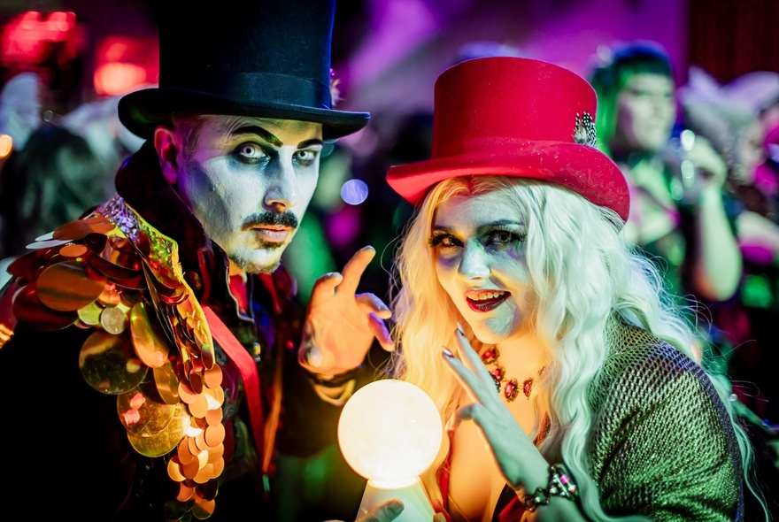 Two people in elaborate fantasy costumes and heavy makeup with an illuminated crystal ball between them. 