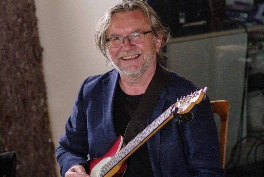 A man with longer grey hair and a short beard smiling broadly while holding a red electric guitar.