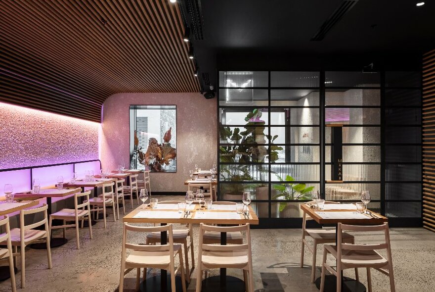 Interior of restaurant with blond wood tables and chairs, large glass doors and mood lighting.