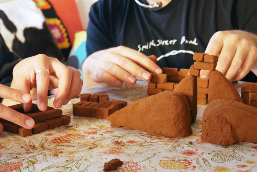 Hands making structures from dark clay at a craft table.