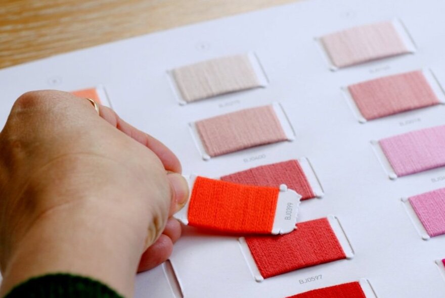 Hand resting an orange colour swag next to rows of reddish-hued fabric samples.