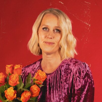 Comedian Claire Hooper wearing a purple dress, she has shoulder length blonde hair, smiles slightly at the camera and holds a bunch of orange roses.