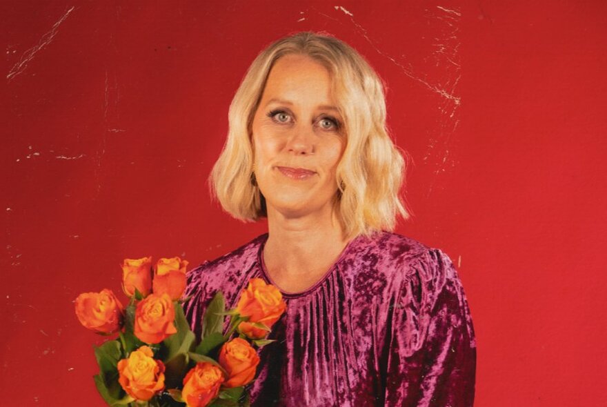 Comedian Claire Hooper wearing a purple dress, she has shoulder length blonde hair, smiles slightly at the camera and holds a bunch of orange roses.
