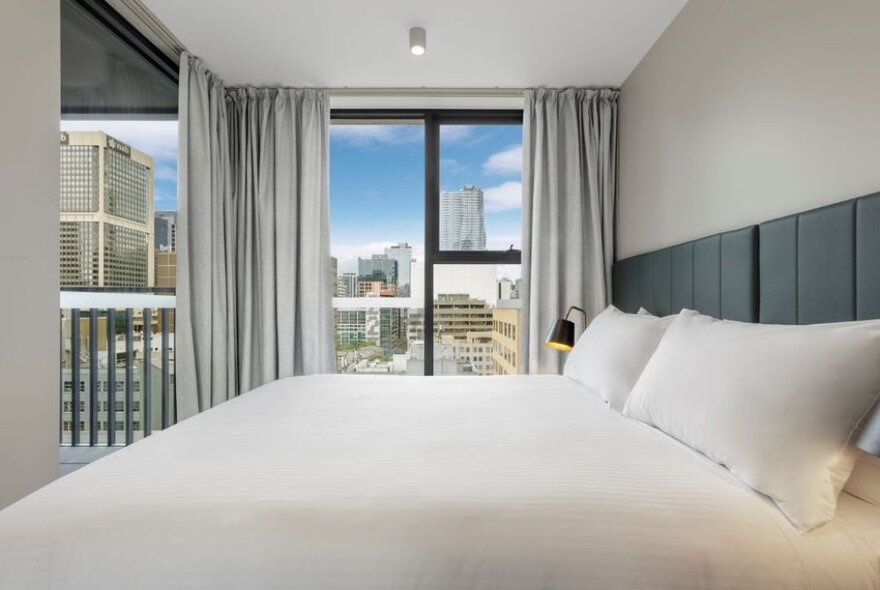 Hotel bedroom with large bed, balcony and expansive windows with city views.