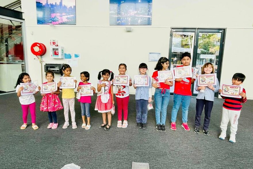 Row of children standing in a line in a room and holding up artworks they've created.