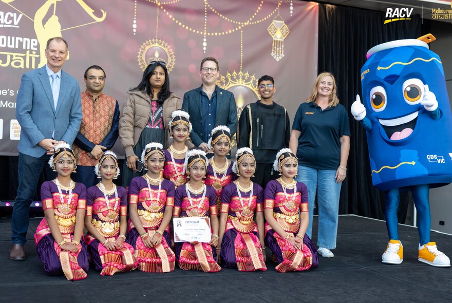 Children in traditional dress kneeling on a stage with adults in front of a large screen.