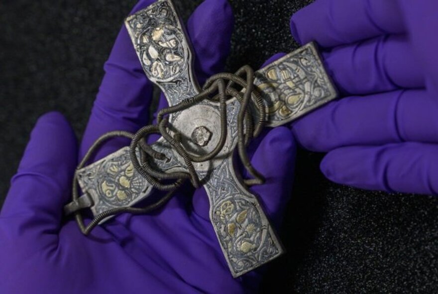 A Viking relic in the shape of a cross in hands wearing royal purple gloves. 