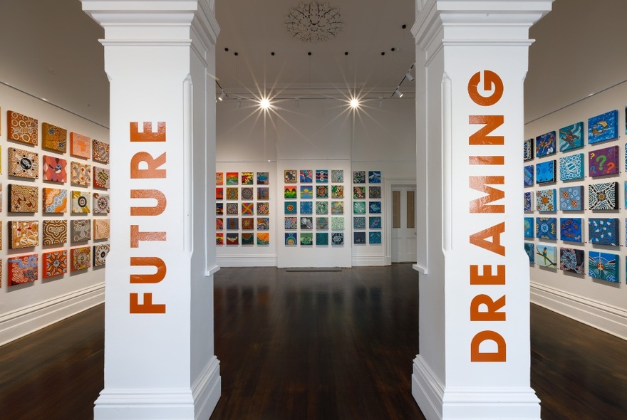 An installation view of many artworks displayed on three walls in a gallery space, with the title of the exhibition FUTURE DREAMING, written on two pillars at the entrance to the exhibition space. 