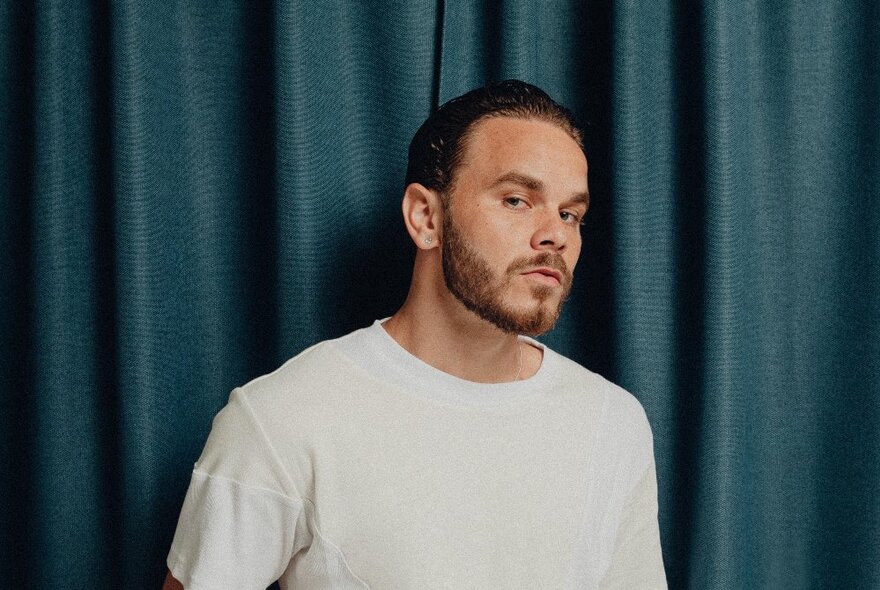 A young man with short cropped hair and a trimmed beard glancing at the viewer, standing in front of a blue curtain.