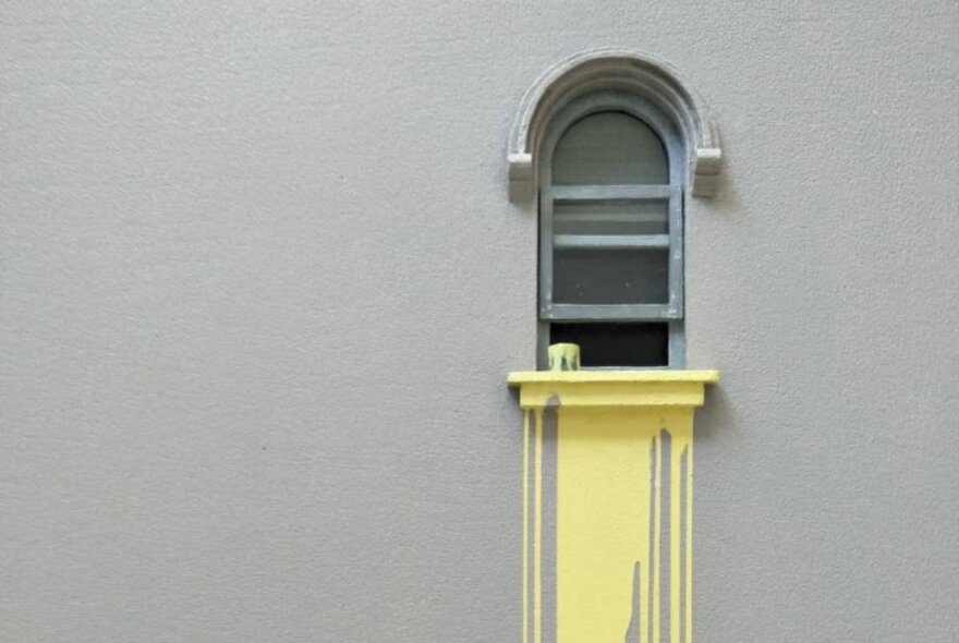 A grey wall and open window and ledge of a building, with a can of paint resting on the ledge and yellow paint dripping down the wall under the ledge.