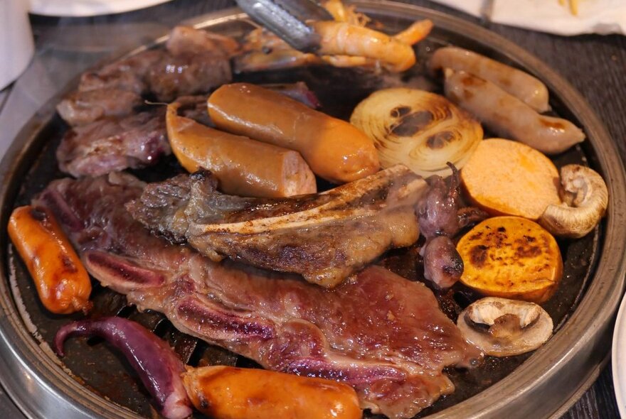 Close-up of a Korean BBQ grill with a large assortment of meats and vegetables: sausages, steak, mushrooms, octopus, sweet potato, onion and prawn.