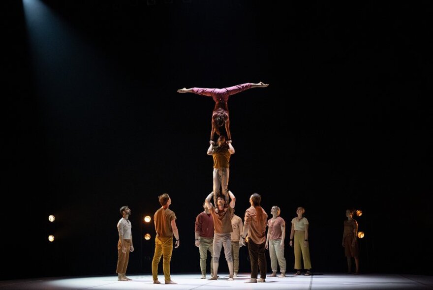 Circus performers on a stage-lit platform standing in a circle around three performers, one standing on another's shoulders whilst holding another one's hands as they balance in a handstand.