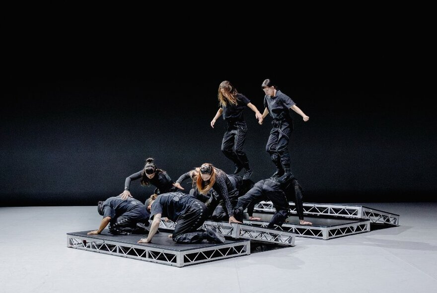 Dancers dressed in dark camouflage pants and shirts posed on three small rectangular raised platforms in a large studio space; white concrete floor and black walls.