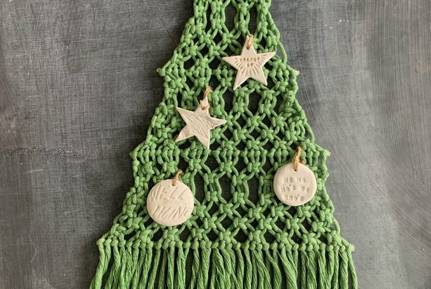 A flat green macrame Christmas tree with small clay decoration on it, hanging on a wall.