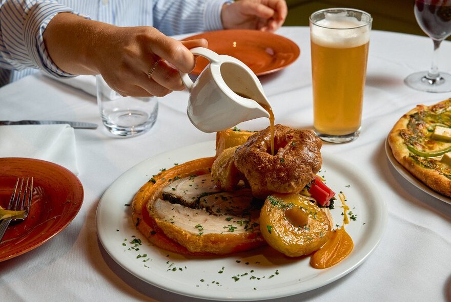 Someone pouring gravy into a Yorskhire pudding on a plate with roast pork and vegetables.