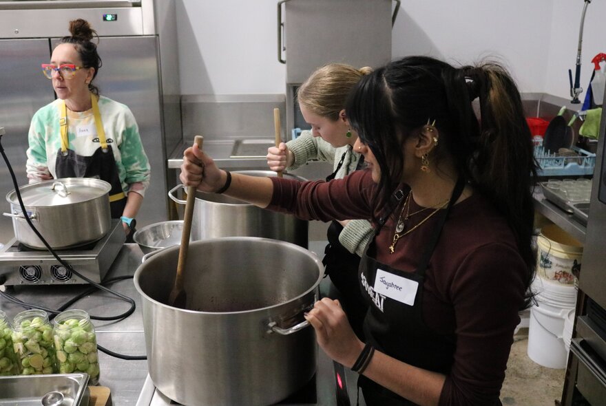 Cooking workshop with people stirring large pots on portable stoves in a kitchen setting.