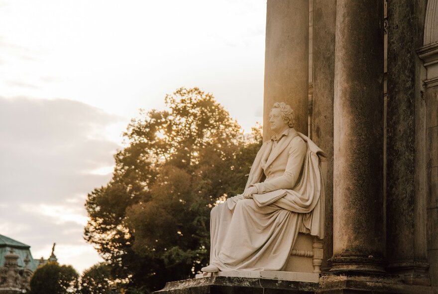 Classically robed statue seated against large pillars with trees in the background.