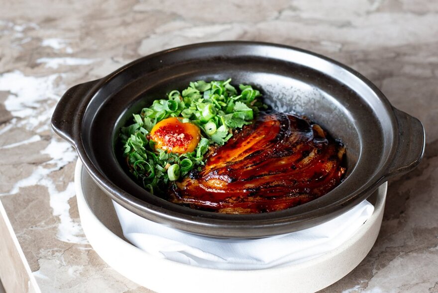 A shallow black dish of grilled meats and greens with an egg yolk, resting on a marble countertop.