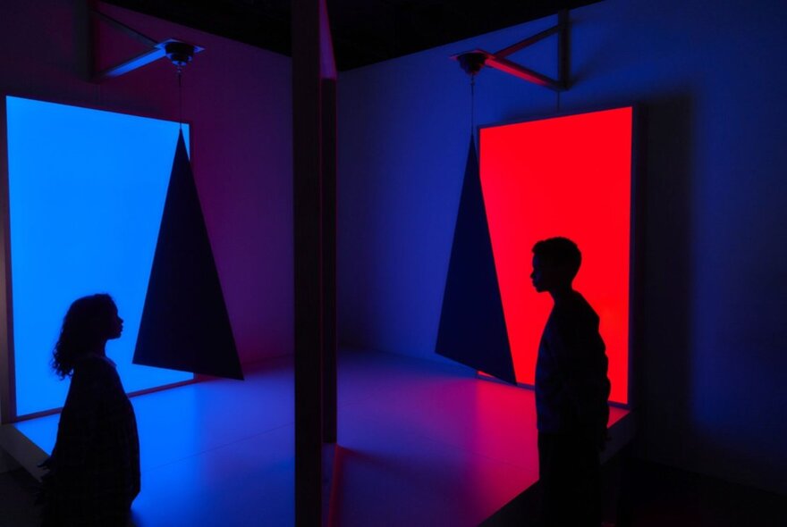 Dark silhouettes against panels of red and blue in a dark exhibition space.