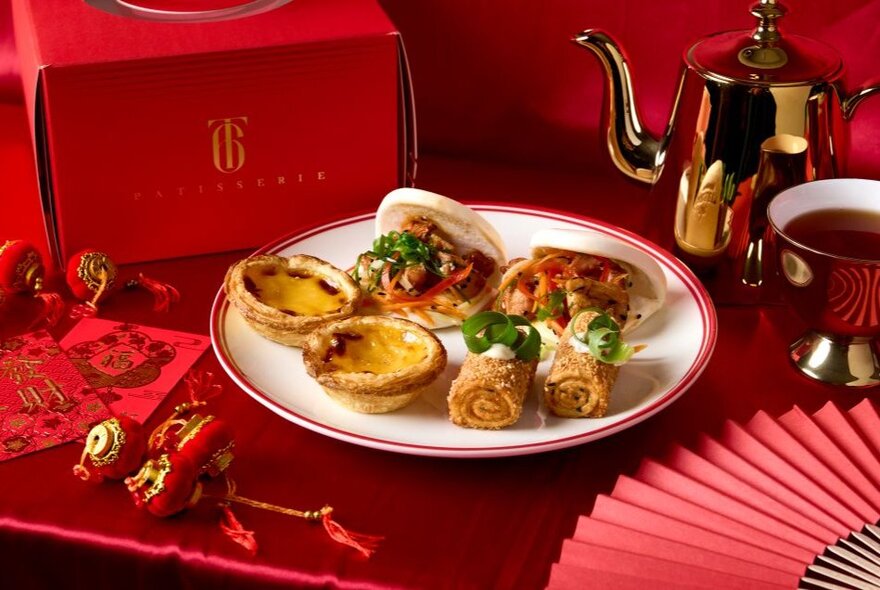 Afternoon tea selection of custard tarts and bao buns on a plate against Chinese New Year red background with fan and ornaments.