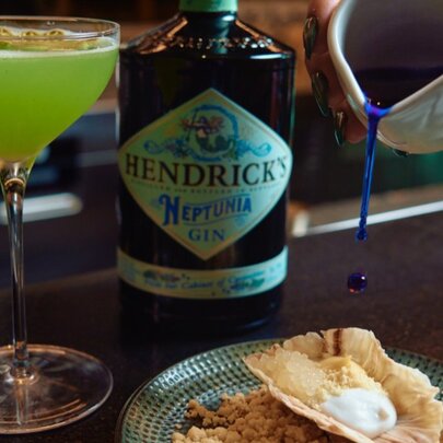 A bottle of Hendrick's Gin next to a lime green cocktail and a hand pouring a blue liquid onto a decadent dessert.