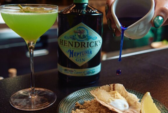 A bottle of Hendrick's Gin next to a lime green cocktail and a hand pouring a blue liquid onto a decadent dessert.