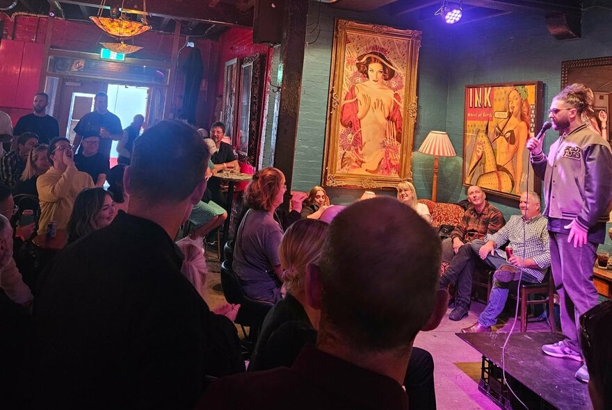 A person holding a microphone performing stand-up comedy on a small stage at Fad Gallery Bar, in front of a live audience.