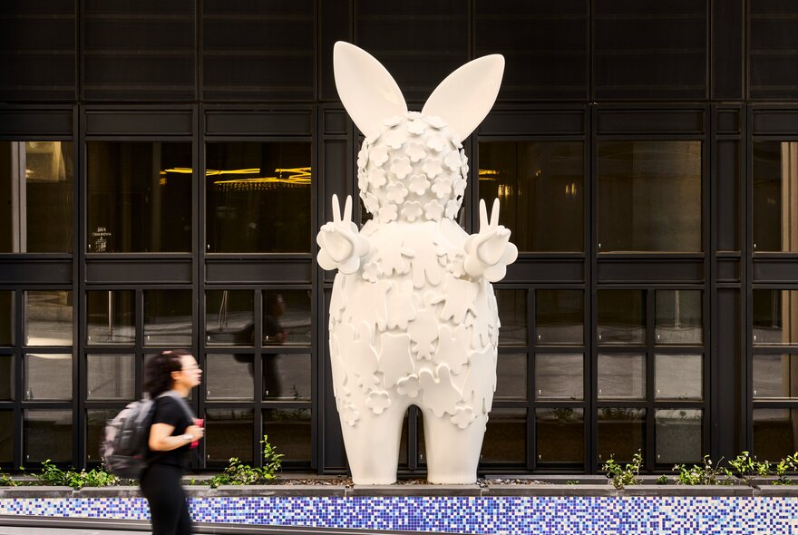 Large white standing rabbit-shaped public artwork, in front of a building, with a person walking past.