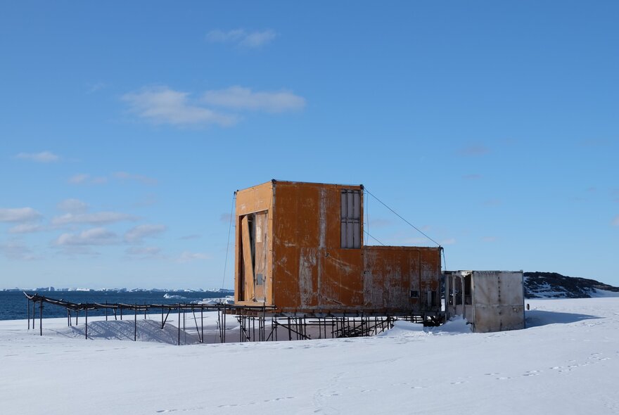 A small, orange-brown research hut on stilts in the snowy, icy landscape of Antarctica, by the ocean under a blue sky.