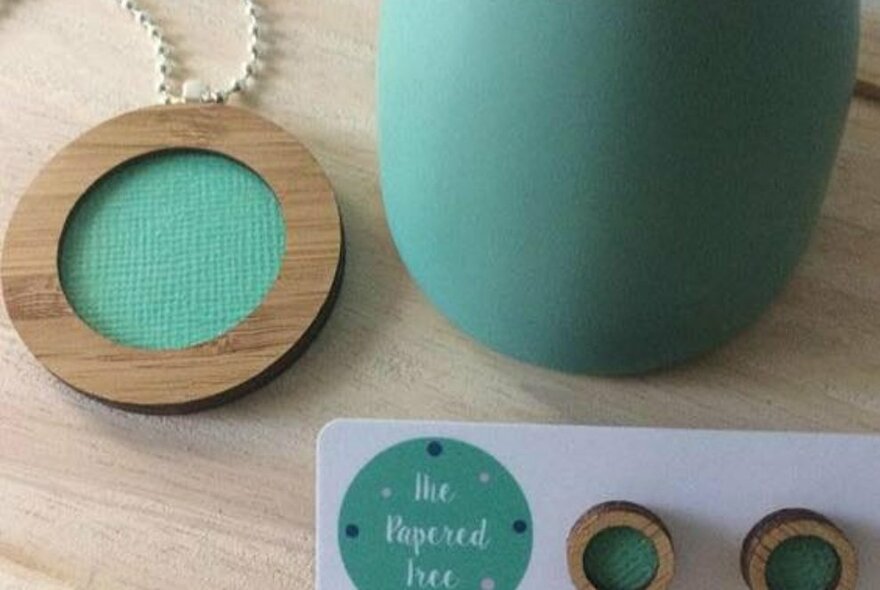 Hand-made teal-coloured paper and wooden jewellery with a teal vase on a wooden surface. 