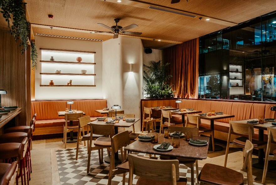 Interior of a restaurant and bar with warm lamp lighting on the wall, arrangements of small tables and chairs, and a patterned black and white check floor.