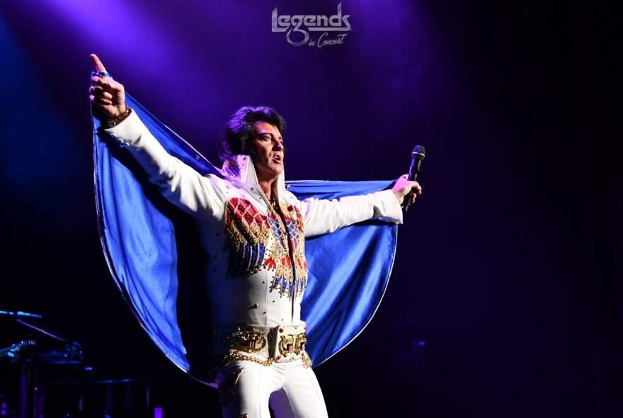 A tribute perfomer on stage dressed as Elvis in a white jumpsuit with blue cape.