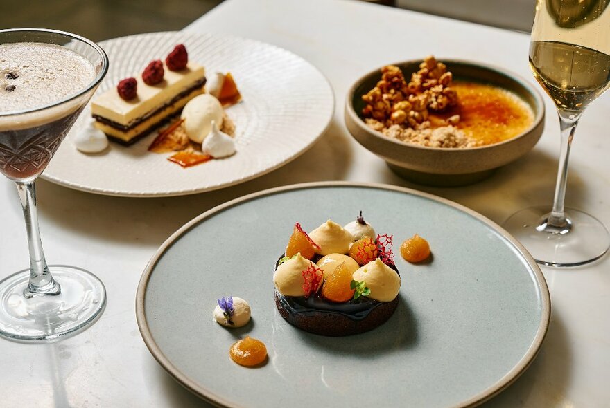 Three plates of desserts, including creme brulee and tiered cake, next to a glass of white wine and cocktail glass with espresso martini.