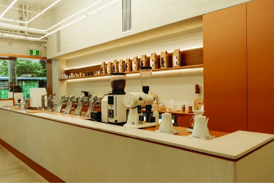 A modern, minimalist well-lit coffee shop interior with a long counter, coffee machines, and bags of coffee beans displayed on shelves.