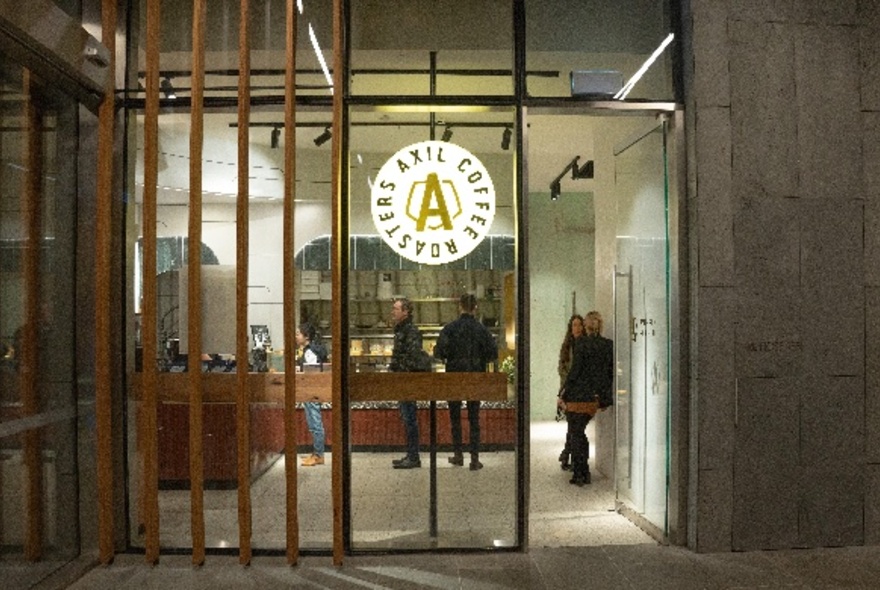 Exterior glass entrance wall of Axil Coffee at Collins Arch, with people standing inside waiting to order coffee.
