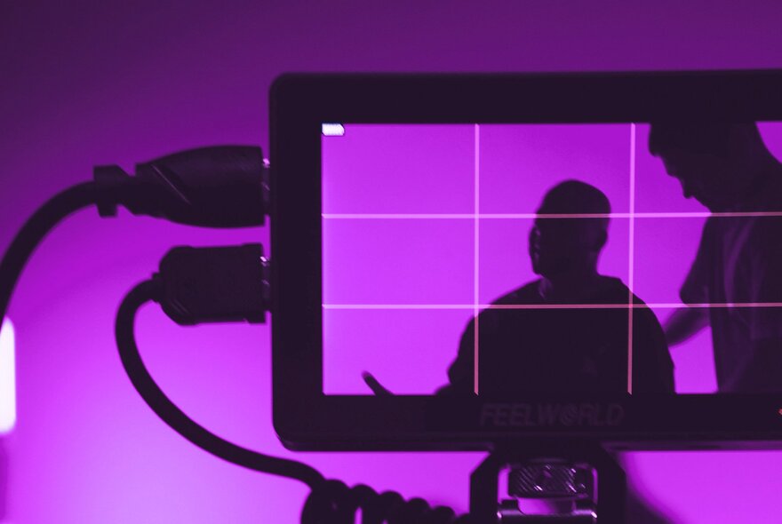 A camera monitor displays the silhouettes of two people against a bright purple backdrop, with a composition grid overlay.