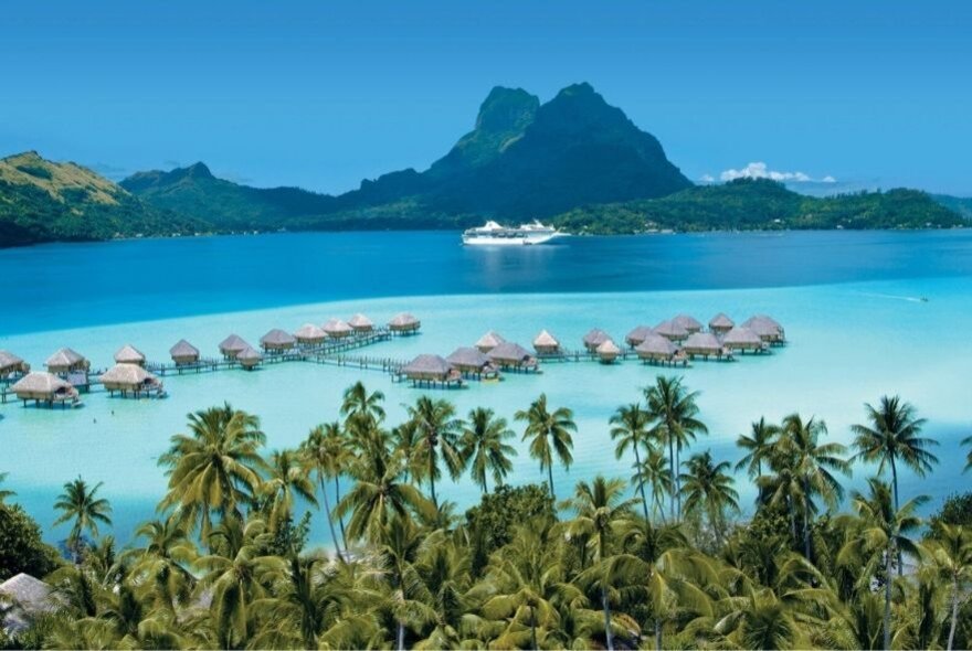 Thatched cabins in a blue sea surrounded by palms and mountains.
