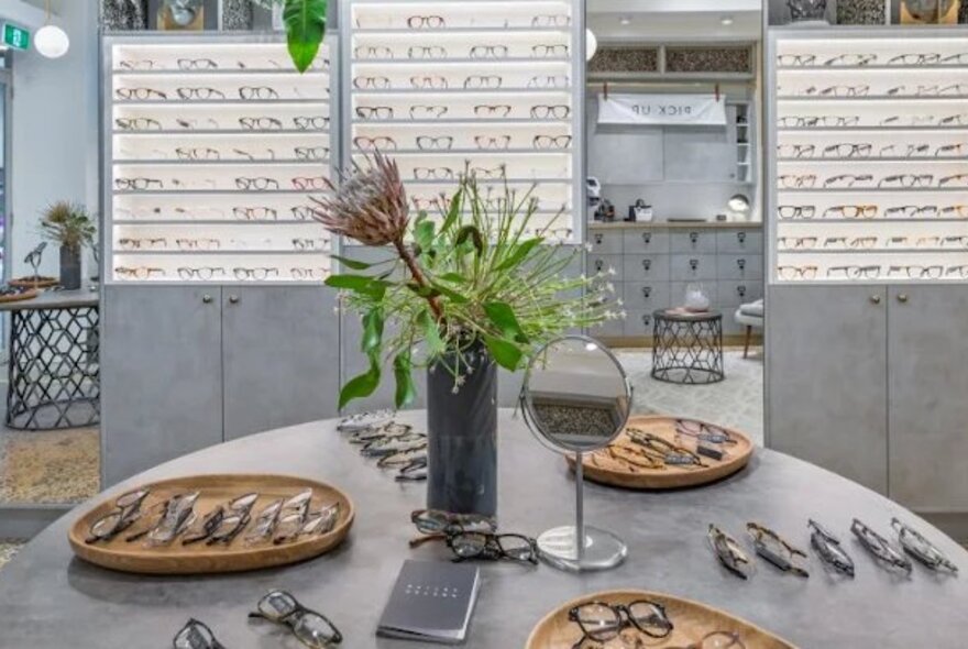 The interior of an eyeglass store, with shelves of eyeglasses along the back wall and a round table in the foreground with glasses laid out on trays and a floral centerpiece.