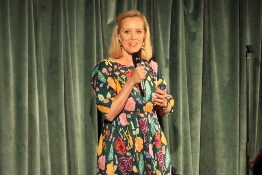The comedian Claire Hooper talking into a microphone they hold in their hand, while standing on a stage in front of a green velvet curtain.