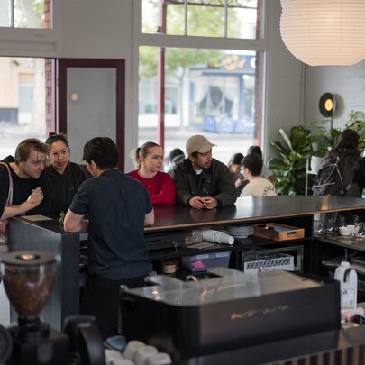 The interior of a coffee shop, seen from behind the counter, with groups of people seated at a bench table waiting for their orders.