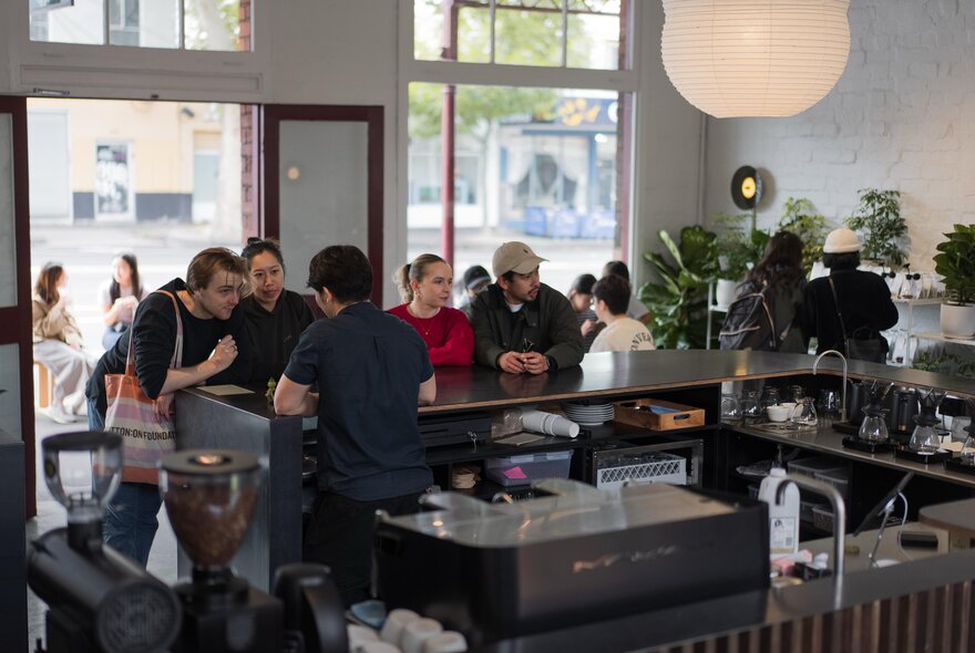 The interior of a coffee shop, seen from behind the counter, with groups of people seated at a bench table waiting for their orders.