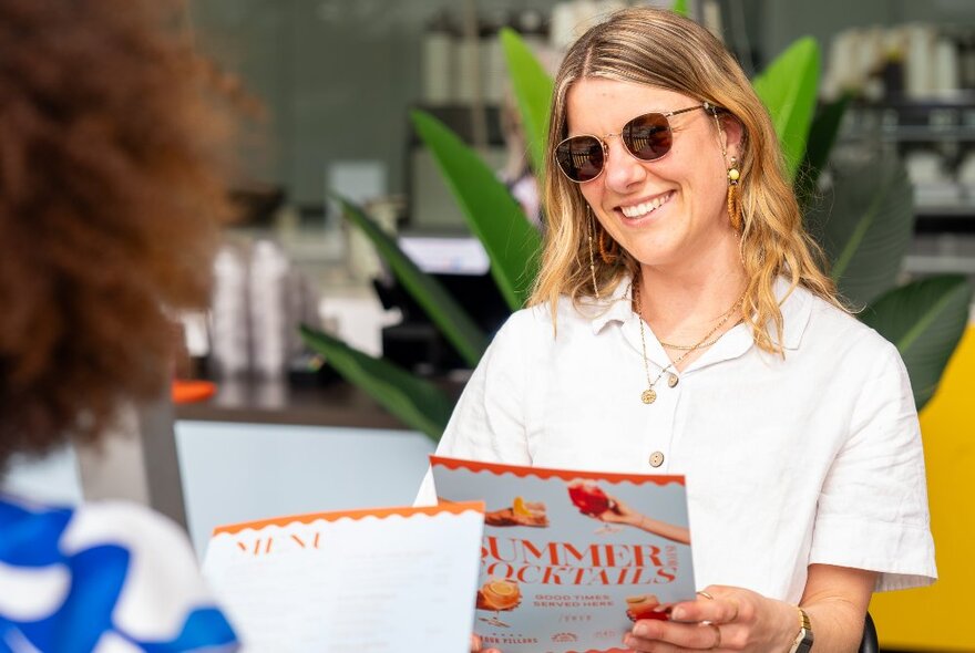 A woman in white shirt and sunglasses, reading a menu at a cafe with the spiky leaves of a green plant behind her.