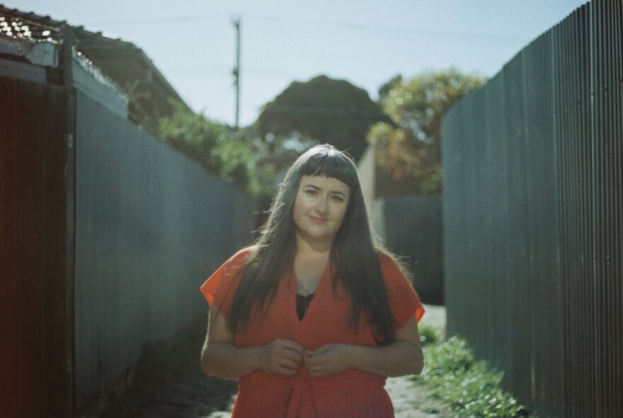 Portrait of a young woman with long brown hair and an orange sleeveless shirt, standing between two tall dark fences.